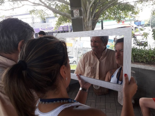 Cláudia está segurando um espelho. Ao lado dela há um homem idoso. No reflexo do espelho dá para ver o rosto dos dois fazendo careta.
